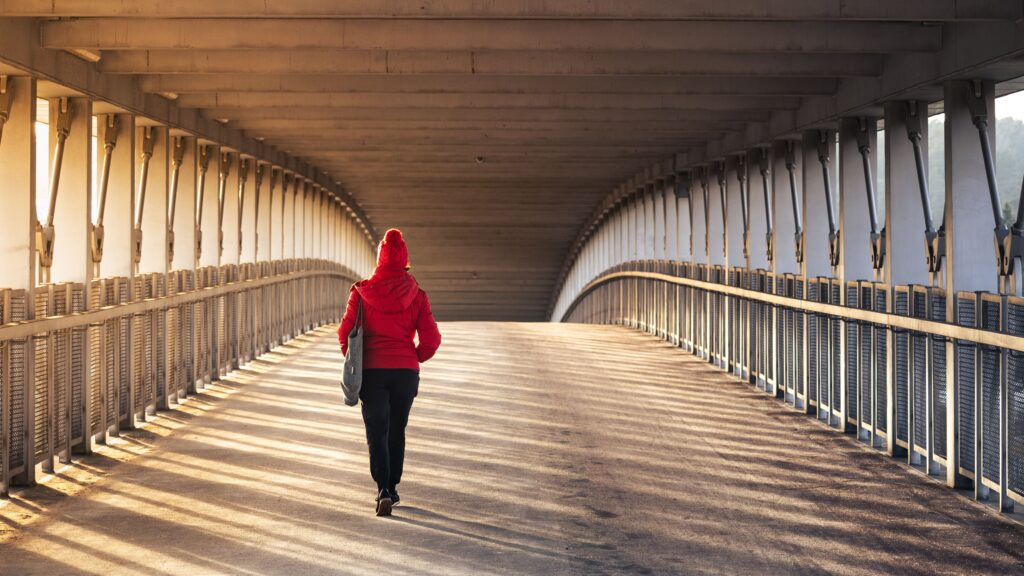 A woman in a red coat walks across a modern bridge, with sunlight casting long shadows.