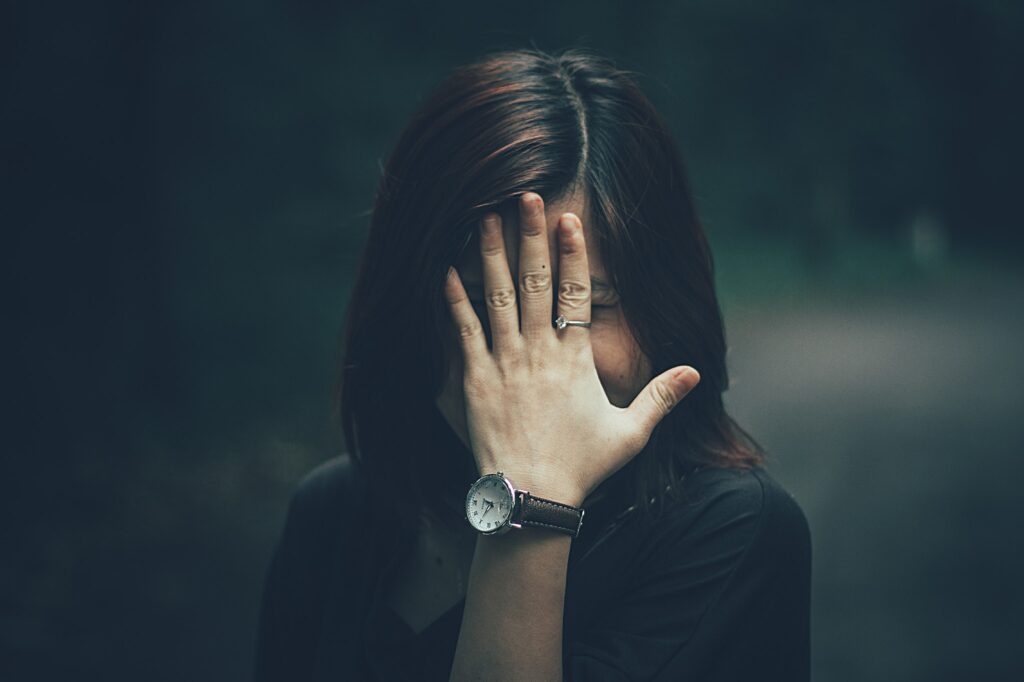 A woman with a wristwatch hides her face with her hand in a moody outdoor setting.