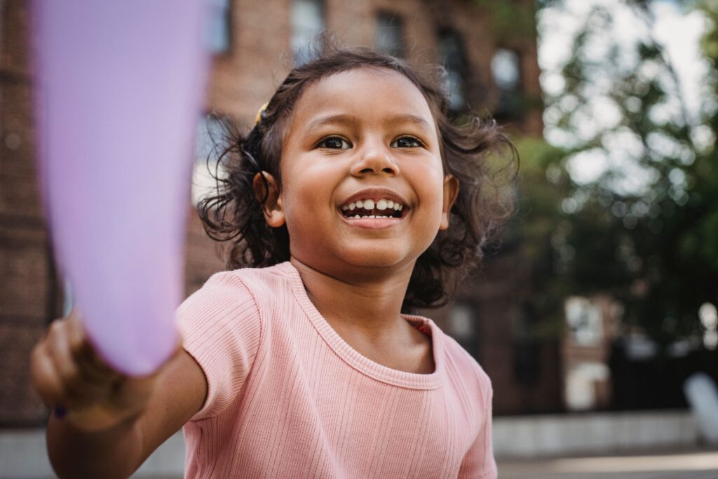 A happy little girl with long hair smiling brightly while playing with a toy outdoors.