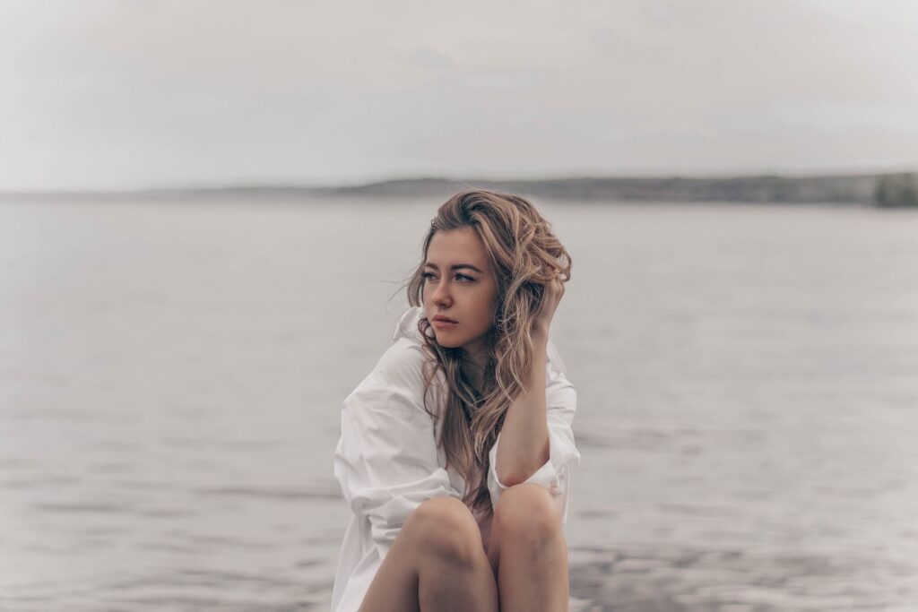 A woman in a white shirt sits by a calm lake, lost in thought.