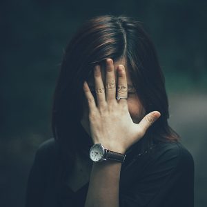 A woman with a wristwatch hides her face with her hand in a moody outdoor setting.