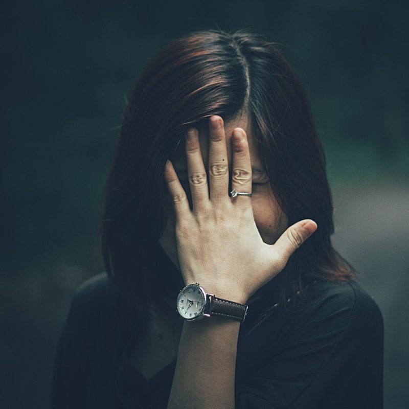A woman with a wristwatch hides her face with her hand in a moody outdoor setting.