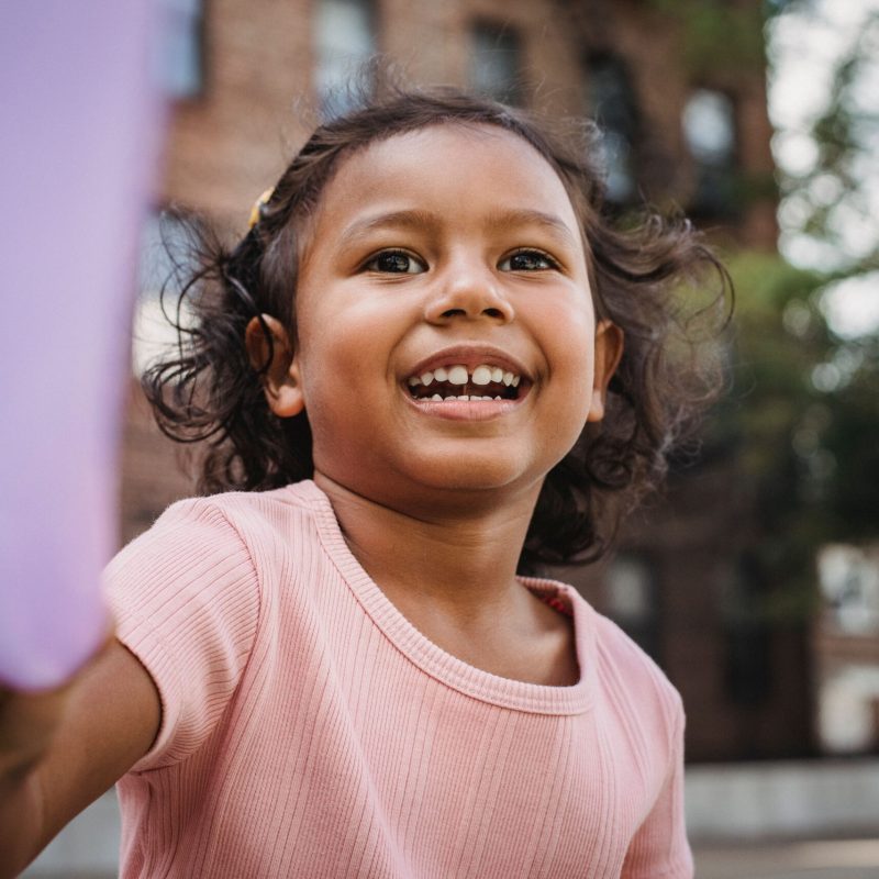 A happy little girl with long hair smiling brightly while playing with a toy outdoors.