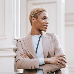 Young dreamy ethnic female executive in formal wear with crossed arms looking away near ornamental column in city